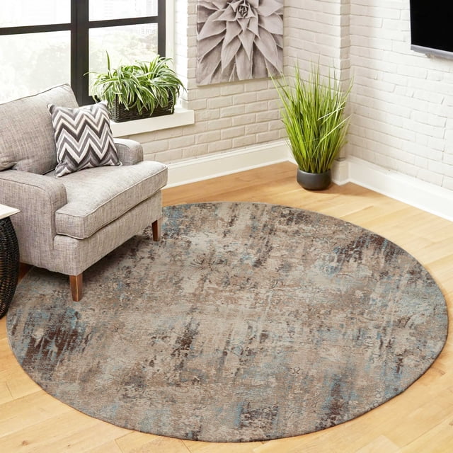 ECARPET Malia Traditional Area Rug for Living Room, Bedroom, Dining Room, Entrance, Hallway and More, round with muted beige, brown, and blue Turkish-inspired patterns on synthetic material, placed on light hardwood floor beneath a gray armchair with a chevron-patterned pillow, next to white brick walls and green plants in a bright room, suitable for high-traffic areas.