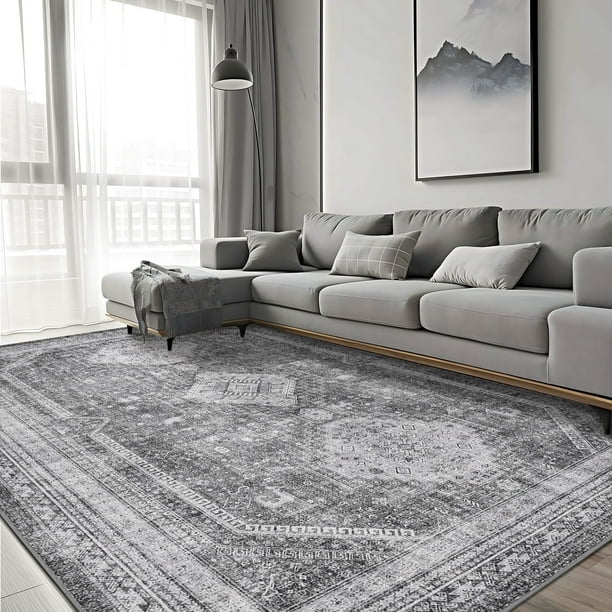 KUETH 5'x7' area rug displayed in a modern living room with a grey sectional sofa and a large window. The ultra-thin, machine washable rug features a vintage-inspired blue and grey abstract design with a low-profile surface that fits seamlessly under furniture. Its non-slip rubber backing ensures stability on hardwood floors, making it ideal for high-traffic areas like living rooms, bedrooms, or dining rooms.