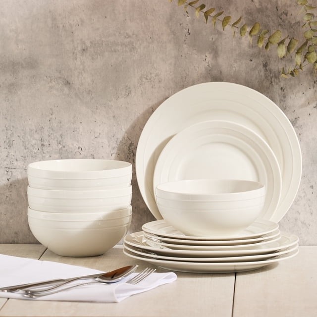 Home Trends Ensemble de Vaisselle d'Environ 12 Pcs en Porcelaine Osseuse Blanche Neuve, Incluant Assiettes à Souper, Assiettes à Salade et Bols avec accents d'Anneaux Concentriques, Disposés sur une surface en Bois Clair avec des Couverts en Acier Inoxydable sur une Serviette Blanche, Sertis contre un Mur Gris Texturé avec des Brindilles d'Eucalyptus, Idéal pour les Repas Quotidiens et les Réceptions Décontractées.