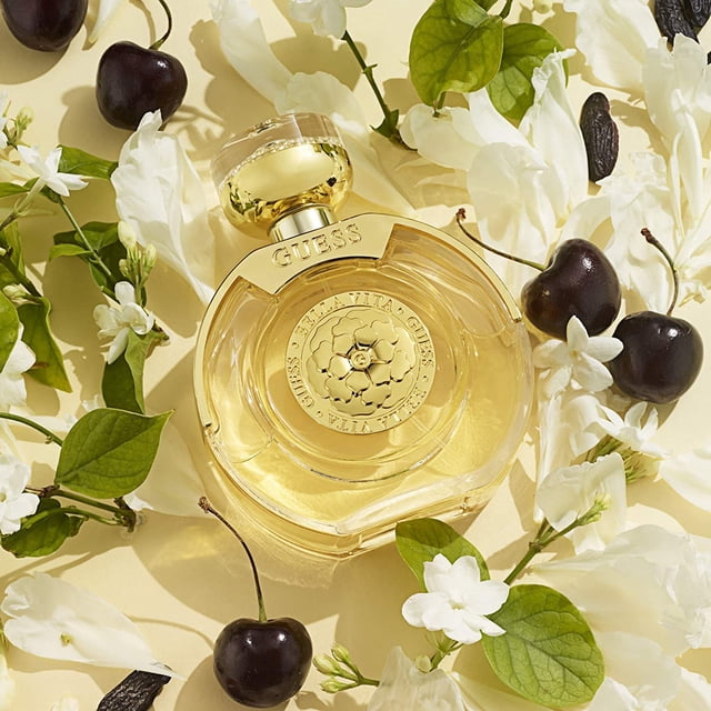 GUESS Bella Vita Fragrance Shimmer Mist 8.4 Fl Oz, a luxurious fruity floral oriental fragrance for women, showcased in a round, golden bottle with floral accents. The bottle is surrounded by jasmine flowers, dark cherries, and vanilla pods on a beige background.