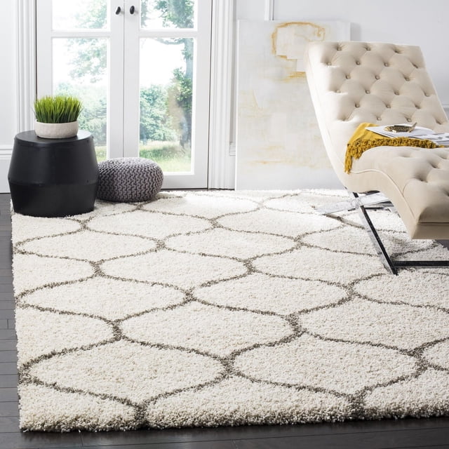 Safavieh Hudson Arline Geometric Shag Area Rug featuring a thick ivory shag pile with gray onion-shaped tile patterns on dark hardwood flooring. Positioned in a modern living room with a beige tufted chaise lounge, black side table with a green plant, and gray knitted pouf near glass doors showing outdoor greenery. Suitable for adding plush texture and durability to high-traffic urban or mod décor spaces.