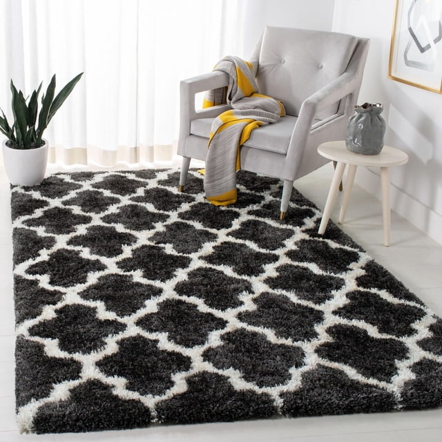 Safavieh Barcelona Anabelle Geometric Shag Area Rug with black and white Moroccan quatrefoil pattern, placed in a bright room with a light grey armchair draped with a grey and yellow throw, a small round wooden side table with a grey ceramic vase, and a green potted plant by sheer white curtains, suitable for adding plush texture and Mediterranean style to accent rug decor.