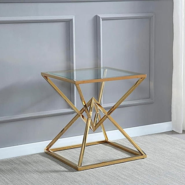 Heavenly Collection Cinna End Gold Table with geometric gold stainless steel frame featuring interlocking angular forms and an 8mm clear tempered glass top, 24 inches high, 21.5 inches wide and deep, shown indoors on a light carpet against a grey paneled wall, suitable as a patio console, side, or accent table.