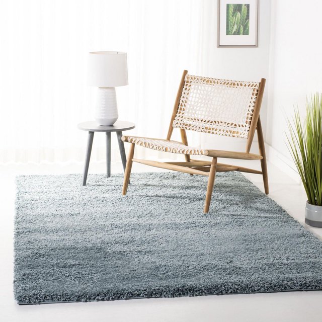 Safavieh California Tapis Shag Uni en Peluche Gris Doux avec un Empilage dense de 4,5 kg par Mètre Carré, Exposé dans une Pièce Minimaliste Lumineuse avec une Chaise Longue en Bois Présentant un Siège et un Dossier Blancs Tissés, une Petite table d'Appoint Ronde Grise avec une Lampe Texturée Blanche, et une Plante Verte en Pot Près d'Un Mur Blanc avec une Oeuvre de cactus Encadrée, Convient pour Ajouter un Confort Moelleux aux Espaces de Vie.