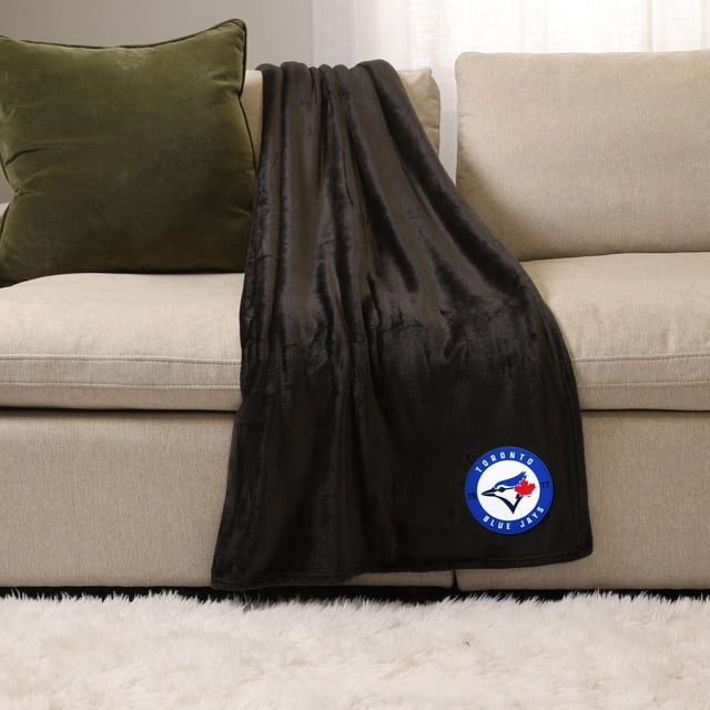 NHL, NBA, MLB Black Sports Throw Blanket, 50" x 60", plush black polyester blanket draped over a beige sofa with a green pillow, featuring a Toronto Blue Jays logo patch in blue, white, and red on the lower corner, suitable for indoor use and cozy warmth.