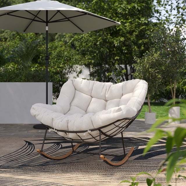 HOMETRENDS Isla Steel Outdoor Rocking Chair with Cushion in cream, featuring a plush, tufted, UV-resistant polyester bouclé cushion tied to a black powder-coated steel frame with woodgrain-finished rocking legs, set on a patterned outdoor rug under a large cream patio umbrella with green foliage in the background.