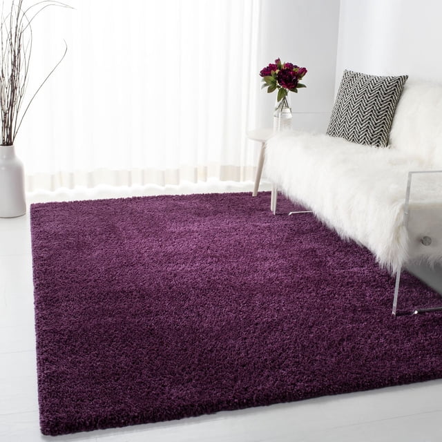 SAFAVIEH August Carlene Tapis Shag en Peluche Uni Violet Foncé sur un Sol Clair, Exposé dans une Pièce Lumineuse avec des Rideaux Blancs Transparents, à Côté d'Un sofa Recouvert de Fausse Fourrure Blanche avec un Coussin en zigzag Noir et Blanc et une table d'Appoint Tenant un vase de Fleurs Rouge Foncé, Souvent Utilisé pour Ajouter du Confort et du style en Peluche aux Espaces de Vie.