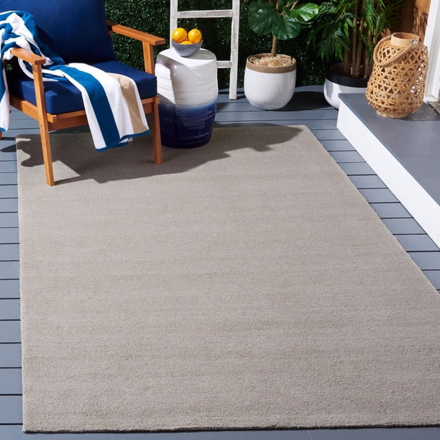 SAFAVIEH Outdoor Micro-Loop Garrett Solid Indoor/Outdoor Area Rug in grey with plush micro-loop pile texture, placed on a grey wooden deck. It pairs with a wooden armchair with navy cushions and a blue and white striped throw, a white and blue ceramic side table holding oranges, and surrounding potted plants and a woven lantern, suitable for outdoor leisure spaces.
