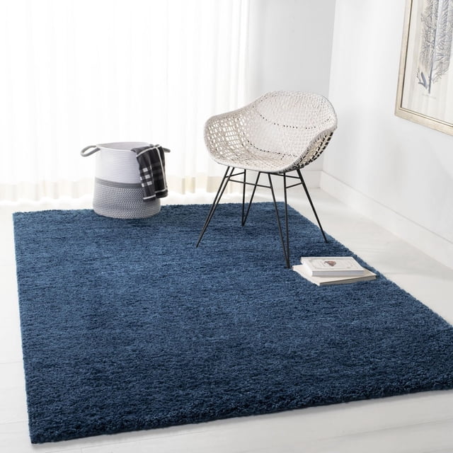SAFAVIEH August Carlene Tapis Shag en Peluche Uni Bleu Foncé, Posé sur un Plancher de Carreaux Blancs dans une Pièce Lumineuse avec des Rideaux Transparents, Présentant une Chaise Tissée Blanche avec des Pattes en Métal Noires et un Panier Gris avec un Tissu Noir et Blanc, Mettant en Valeur la texture de Fil de Polypropylène Peluche Rug's et un style Décontracté de la Côte Ouest, Convenant pour Améliorer le Confort et la Déco dans les Intérieurs de la Maison.