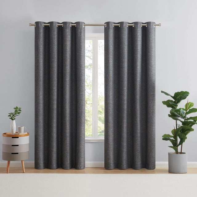 Eclipse Auden 63" grommet blackout window curtains panel in dark grey with textured weave, hanging on a metal rod over a window. Designed for light control, total privacy, and noise reduction. The room has light grey walls, wooden floor, a small round side table with a plant and cup on the left, and a tall potted plant on the right. The curtains are machine washable and suitable for enhancing living spaces.