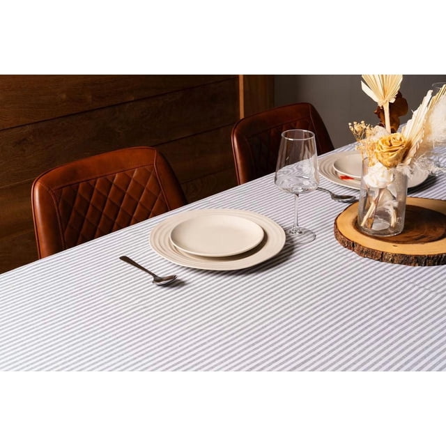 Fabstyles Nappe à Rayures Bistro 100 % Coton Gris avec Motif à Rayures Classique, Couvrant un Ensemble de Table à Manger avec des Assiettes en Céramique Crème, une Cuillère, un Verre à Vin Transparent et un arrangement de Fleurs Séchées dans un vase en Verre sur un Plateau Rond en Bois Naturel, Entourée de Chaises en Cuir Matelassé Brun et d'Un Fond Mural en Bois, Idéale pour les Repas à l'Intérieur et les Événements Spéciaux.