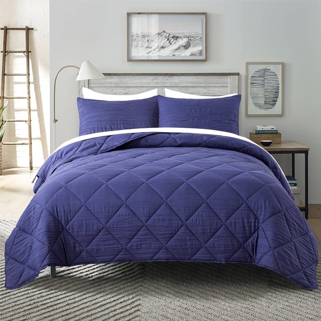 JML 2 Pieces Navy Twin Comforter Set displayed on a bed in a modern bedroom setting. The set includes a quilted navy comforter with square patterns and a matching pillow sham, made of soft 90GSM brushed microfiber and filled with 250GSM polyester for warmth and comfort. This microfiber bedding set features a vintage creased texture and is suitable for all seasons. The room features neutral decor with framed artwork, a wooden nightstand, and a ladder accent piece.