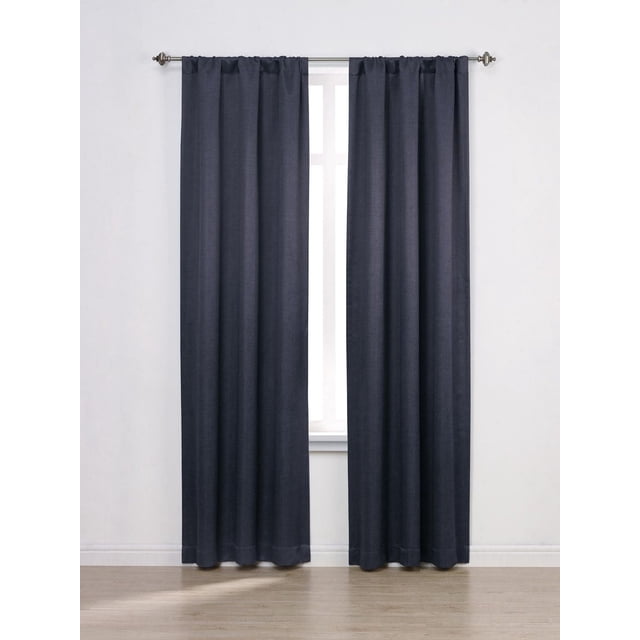 Mainstays Chambray Blackout 63" Window Curtains Panel Pair in dark blue chambray texture with rod pocket design, each panel 40 inches wide and 63 inches long, offering 100% blackout technology for bedroom light control and privacy in a minimalist white room with light wood flooring.