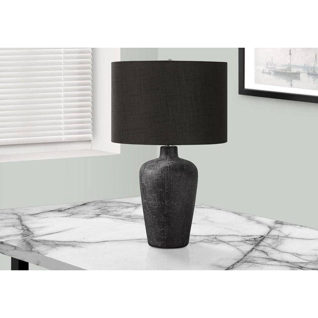 Monarch Specialties Lighting table lamp, 24" tall, with a textured black ceramic vase-shaped base and black fabric drum shade, placed on a white marble end table near a window with white blinds and a framed nautical print on a pale grey wall, ideal for living rooms, bedrooms, or offices.