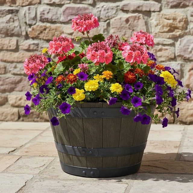 Hometrends 19in Whiskey Barrel Resin Planter, Brown, a round resin planter with wood grain texture and black metal bands, filled with pink geraniums, yellow and orange marigolds, and purple petunias, placed on a stone patio against a rustic stone wall, ideal for outdoor garden decoration.