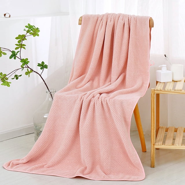 Large bath towel in a soft pink color, made of cotton with a textured pattern, displayed draped over a wooden chair in a bright bathroom setting. The towel measures 55.11x27.55 inches, offering an oversized design suitable for plus-size users. It is highly absorbent, quick-drying, and designed for comfort and practicality. Nearby, a small wooden shelf holds bathroom essentials, complementing the towel's elegant and functional appearance.