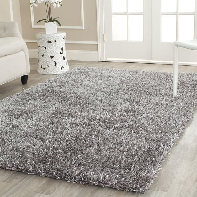 Safavieh New Orleans Darius Solid Shag Area Rug in grey with an extra-high pile of hand-tufted polyester fibers, offering a plush, textured surface on light wood flooring in a neutral-toned room with white French doors, a white ceramic side table, and light furniture. It is also ideal for adding softness to lounge or reading areas.