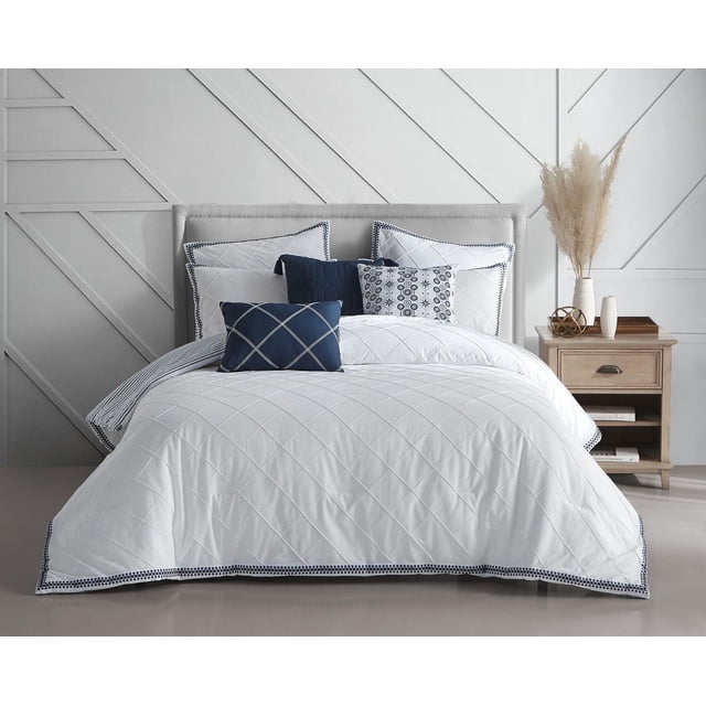 Beco Home Pintuck White 3 Piece Cotton Comforter Set on a bed with a grey upholstered headboard, featuring a white cotton comforter with diamond pintuck stitching and navy embroidered edges. Includes two matching shams with navy trim and decorative navy and white pillows. The bed is styled in a minimalist room with a wooden nightstand holding pampas grass and decor, offering a clean, airy bedding look ideal for a relaxed bedroom setting.