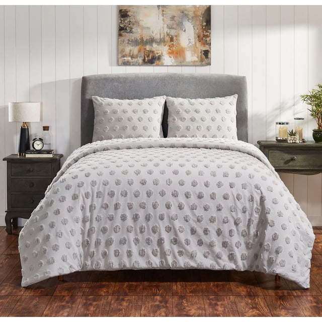 Ensemble de Couette en Coton Venice sur un Lit avec Tête de Lit Rembourrée Grise, Comprenant une Couette en chenille de Coton Capitonnée et Deux Taies Très Grand Size avec un Motif à Pois Capitonnés Gris. l'Ensemble de Literie 100 % Coton Est Conçu dans une Chambre avec des Planchers en Bois, des Murs à Panneaux Blancs, une table de Chevet en Bois Foncé avec une Lampe et une Horloge d'Un Côté, et une table d'Appoint Assortie avec des Bougies et une Plante de l'Autre, Accentuée d'Un Tableau Mural Abstrait.