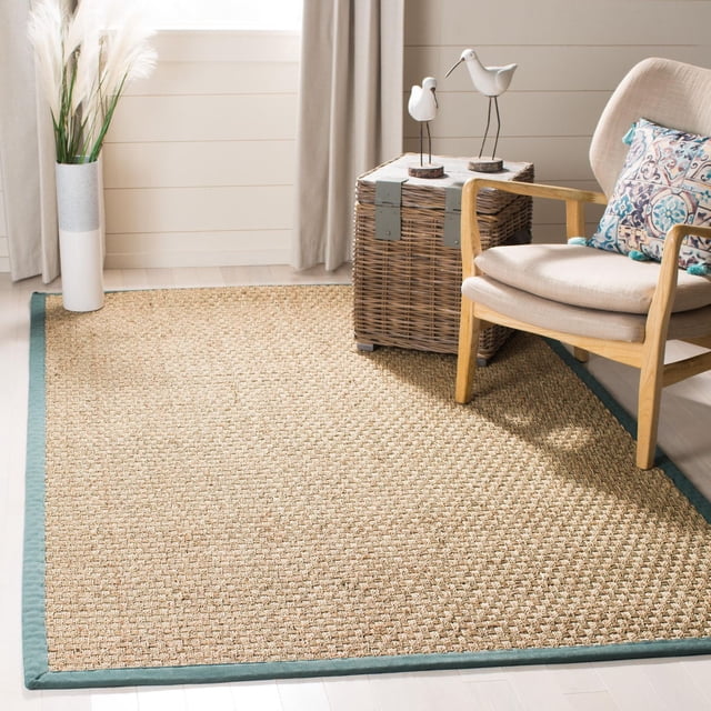 Safavieh Natural Fiber Arbor Border Area Rug featuring woven natural sisal texture with a teal fabric border, placed on light hardwood flooring beside a beige wooden armchair with a patterned cushion and a wicker side table with white bird figurines, suitable for coastal and casual interiors.