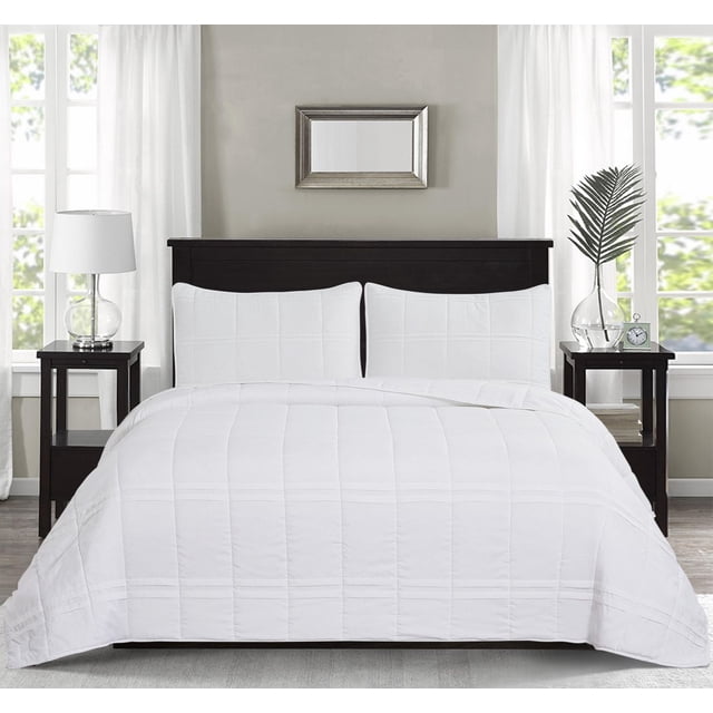 Chaps 3-Piece Jersey Knit Microfiber Quilt and Sham Set - Solid Coverlet Comforter in white displayed on a dark wood bed frame with two matching pillow shams featuring a stitched grid pattern. The bedroom includes dark wood nightstands with a glass lamp and a vase with a palm leaf, framed by white curtains and large windows, suitable for enhancing bedroom bedding decor.