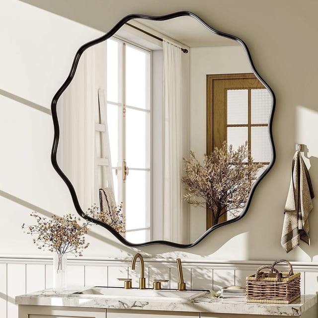 BEAUTYPEAK 30" Wavy Round Bathroom Mirror Wall Decor with Deep Frame, Black. This wall-mounted mirror features a unique wave-shaped edge and a deep black frame, adding a modern and artistic touch to any space. Made with high-definition tempered glass, it provides a clear, distortion-free reflection and enhanced durability. The mirror is displayed in a bathroom setting above a marble countertop with gold fixtures, surrounded by neutral decor and natural light.