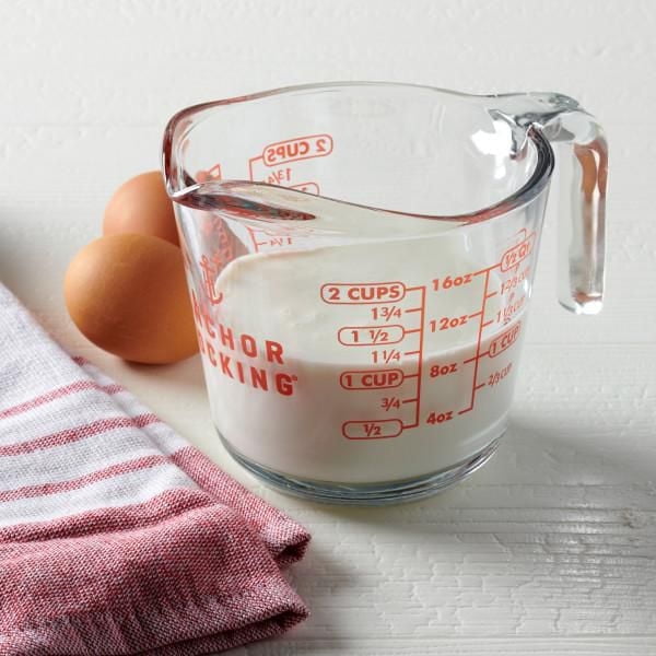 Anchor Hocking 500ml measuring cup made of thick tempered glass with bright red measurement markings in cups, ounces, and millilitres, filled with milk, featuring an easy-pour spout and ergonomic handle, placed on a white surface beside two brown eggs and a red-striped kitchen towel, ideal for precise cooking and baking.