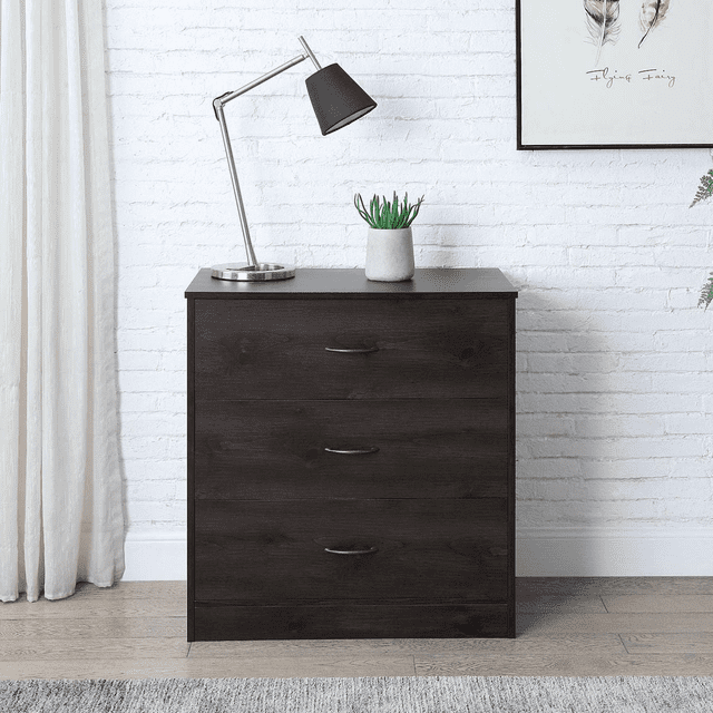 Mainstays 3-Drawer Dresser in dark oak finish with three metal-handled drawers providing ample storage, placed against a white brick wall in a kids' bedroom. The dresser holds a silver desk lamp with a black shade and a small potted succulent, standing on light wood flooring with a grey rug, flanked by white curtains and framed feather artwork.