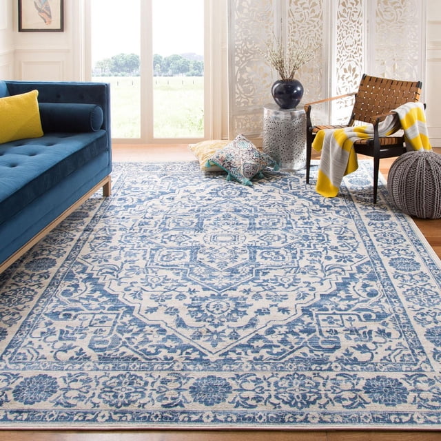 SAFAVIEH Brentwood Jaymz Floral Bordered Area Rug in blue and cream with intricate floral and geometric motifs, placed in a bright living room with a blue velvet sofa and yellow pillow, a woven leather chair draped with a yellow and gray striped throw, floor cushions, a gray knitted pouf, and a round side table with a dark vase and dried branches, suitable as an accent rug for traditional or transitional decor.