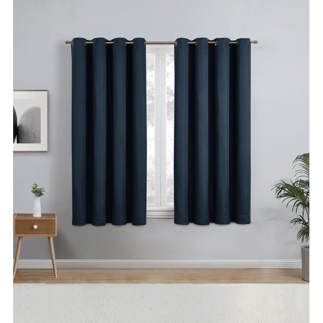Eclipse Heath 63" Grommet Blackout Window Curtains Panel in indigo blue with grommet top hanging on a metal rod, blocking light from a window in a minimalist room with grey walls, wooden floor, a small wooden side table with a white vase and plant, and a potted green plant. Suitable for privacy and energy savings.