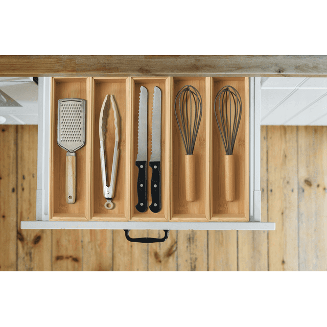 Better Homes & Gardens Bamboo Long Drawer Organizer with five compartments holding a metal grater with wooden handle, metal tongs with silicone tips, two serrated knives with black handles, and two metal whisks with wooden handles in an open white kitchen drawer over a wooden floor, ideal for kitchen utensil organization.