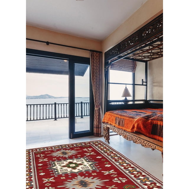 ECARPETGALLERY 100% Wool Traditional Indoor Area Rug with bold geometric Persian Kazak-inspired patterns in red, cream, and green tones, placed on a bedroom floor featuring a carved wooden canopy bed with an orange and brown cover, near glass doors opening to a balcony overlooking the sea and hills, also suitable for entrance, living, or dining rooms.