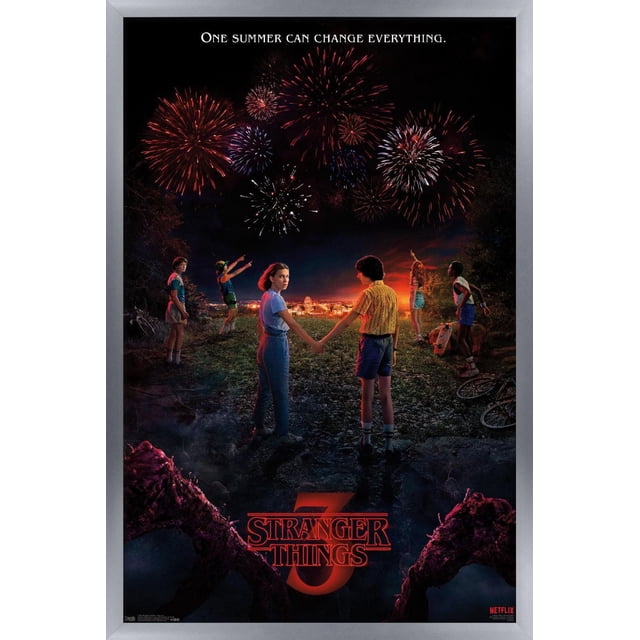 Netflix Stranger Things: Season 3 - Key Art Wall Poster, 22.375" x 34" framed, depicting six children standing on a rocky path at night overlooking a town illuminated by fireworks. Two children hold hands in the center while others point to the sky. The poster includes the tagline 'One summer can change everything' and the red Stranger Things 3 logo, with dark creature limbs framing the bottom edges.