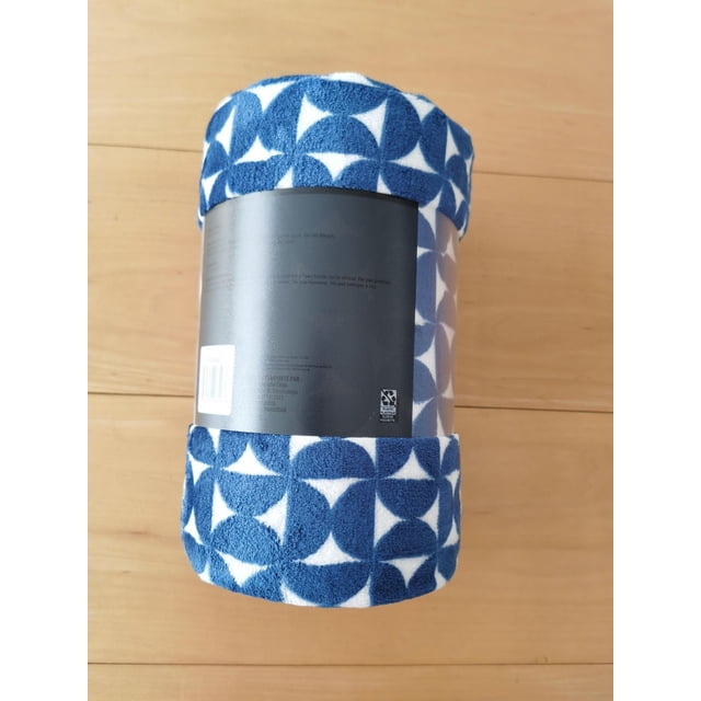 Couverture Jetée en Peluche Roulée avec Étiquette Noire, Présentant un Motif Géométrique Bleu et Blanc en Quart de Cercle, Convient comme Accessoire de Literie Ou Jeté Douillet, Exposée sur une surface en Bois Clair.