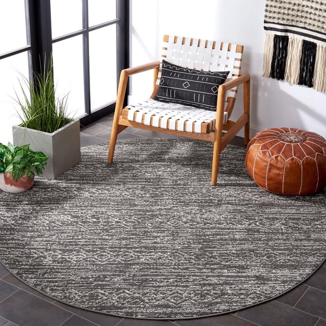 SAFAVIEH Cottage Alease Tapis Géométrique d'Intérieur/extérieur en Gris avec un Motif Géométrique, Tissé Mécaniquement à Partir de Fibres Synthétiques Durables. le Tapis Rond Est Posé sur un Plancher de Tuiles Foncées dans une Pièce Lumineuse avec un Fauteuil en Bois Présentant des Sangles Tissées Blanches et un Coussin Géométrique Noir, un pouf en Cuir Brun, et Deux Plantes en Pot, Convient à une Utilisation Intérieure Ou Extérieure à Fort Achalandage.