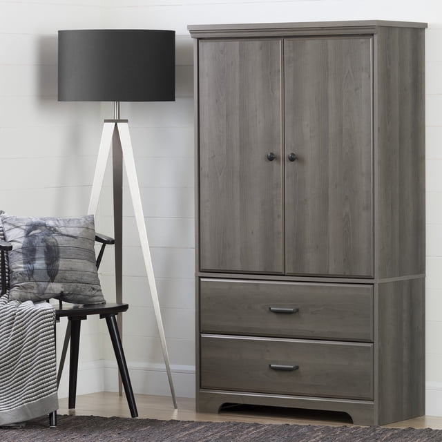 South Shore Versa 2-Door Armoire with Drawers in gray maple finish, featuring two doors with round black knobs, two spacious drawers with rectangular handles, top molding, and curved cutout base, shown in a neutral room beside a black tripod floor lamp and a chair with a bird-patterned pillow and gray throw, ideal for bedroom or living room storage.