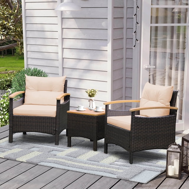 Costway Patiojoy 3PCS Patio Rattan Furniture Set with two mix brown PE rattan chairs featuring beige cushions, curved wooden armrests, and a coffee table with an acacia wood tabletop. This outdoor conversation set is displayed on a grey rug in a patio setting with a white wooden wall and surrounding greenery.