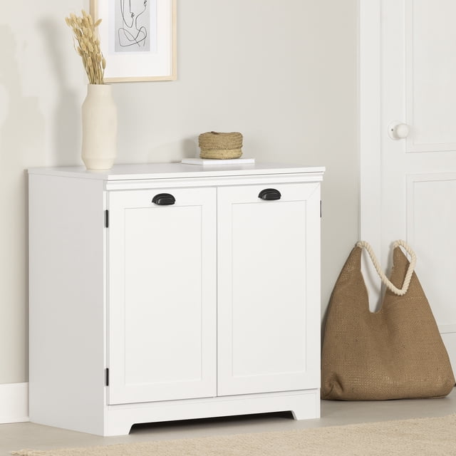 South Shore Prairie 2-Door Storage Cabinet in pure white with framed doors and black metal handles, designed for entryway use with two storage compartments and an adjustable shelf, shown against a beige wall with a vase of dried flowers and a woven basket on top, beside a beige woven tote bag on the floor.