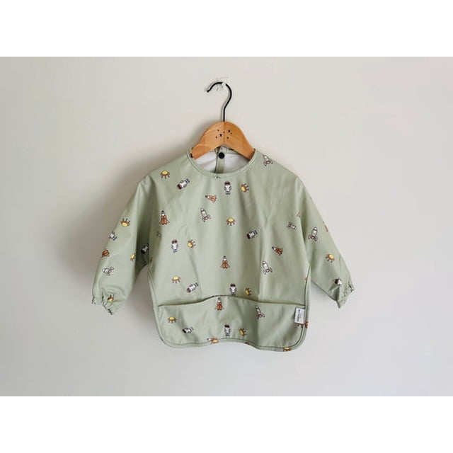 Waterproof long sleeve baby toddler feeding bib with a rockets print, designed for ages 6 to 30 months. Made from 100% polyester with OEKO-TEX certified fabric, it features snap buttons for easy wear and a mess-proof design. The bib is light green with colourful rocket and space-themed illustrations, offering full coverage with elasticated cuffs and a front pocket. Displayed on a wooden hanger against a plain wall.