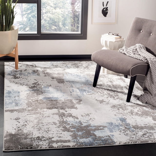 SAFAVIEH Craft Constantine Abstract Area Rug 5'3"x7'6" with a muted abstract pattern in grey, blue, and beige tones and subtle etched pile texture. Displayed in a contemporary room with a grey upholstered chair, knitted throw, white side table with books and glasses, near a large window and potted plant on a dark floor. Ideal for adding modern style and comfort to living areas.