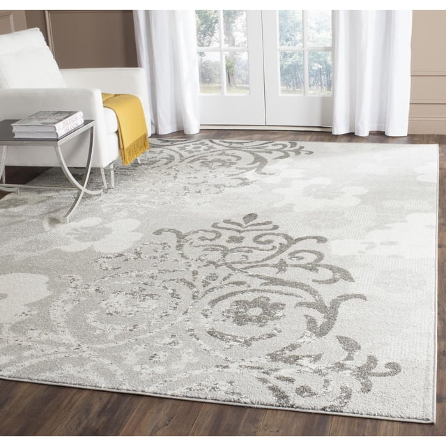 SAFAVIEH Adirondack Hortense Floral Area Rug in a living room, featuring a rectangular polypropylene rug with a muted grey and cream floral scroll pattern, placed on dark hardwood flooring near a white armchair with a yellow throw and white curtains over glass doors, suitable for adding rustic lodge style to home decor.