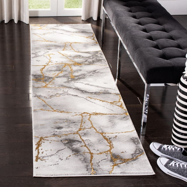 SAFAVIEH Craft Paul Abstract Marble Area Rug runner with white background and gray and gold swirling marble patterns, made of soft synthetic yarns for durability and comfort, placed on dark hardwood floor beside a black tufted bench with chrome legs, suitable for contemporary hallway or entryway decor.
