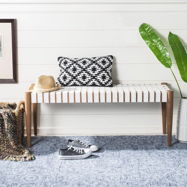 Safavieh Amalia Leather Bench with honey finished mindi wood legs and woven cognac leather seat, styled with a black and white geometric pillow and straw hat, placed against a white shiplap wall on a blue textured rug with a woven basket and green plant, suitable for entryway or bedroom seating.
