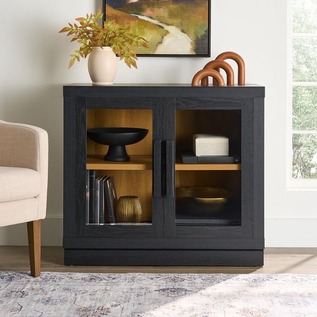 Better Homes & Gardens Ezra Storage Cabinet in black wood with two framed, safety-tempered glass doors and black handles, featuring an adjustable natural wood shelf inside. The cabinet displays a black pedestal bowl, books, a gold vase, a black tray with a beige box, and a dark bowl. Positioned on a light wood floor beside a beige chair, topped with a cream vase holding green and orange leaves and a copper decorative sculpture, against a white wall with a landscape painting and a window showing greenery.