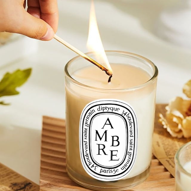 Diptyque Ambre Diffuser featuring a glass container with a warm amber-colored wax and a lit wick, exuding a luxurious and natural amber aroma. The label on the front displays 'Ambre' in bold text, surrounded by an elegant oval design. The diffuser is placed on a wooden surface with decorative elements like dried flowers in the background, creating a cozy and sophisticated ambiance.
