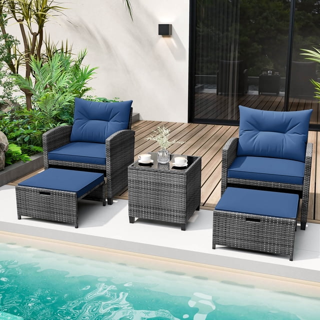 Costway 5 Piece Patio Rattan Furniture set featuring two navy-cushioned chairs, two versatile ottomans, and a tempered glass coffee table. The mix gray PE rattan construction is durable and fade-resistant, with ergonomic backrests and curved armrests for comfort. The ottomans can serve as footrests, extra seating, or side tables. The coffee table includes a tempered glass top secured with suction cups. Set displayed poolside with lush greenery and a wooden deck in the background.
