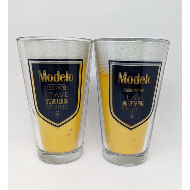 Modelo Verres Pinte Salute To Veterans - Ensemble de 2, Présentant Deux Verres à Bière de 16 Onces avec le logo Modelo et le Texte « Proud Partner Leave No Veteran Behind » en Or sur Fond Bleu Marine. Parfaits pour les Passionnés de Bière et les collections de Vaisselle de Bar, Ces Verres Honorent les Anciens Combattants.