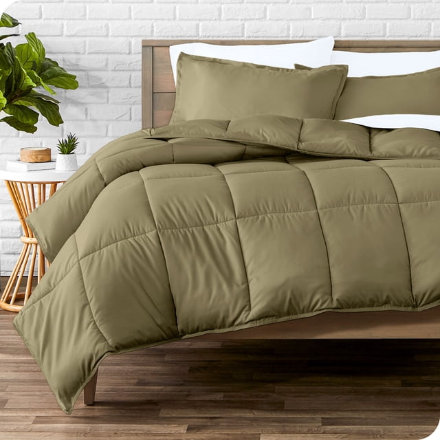 Bare Home Goose Down Alternative Comforter Set - 3 Piece Set - All Seasons - Oversized King, Olive. A front-facing view of the comforter set displayed on a wooden bed frame in a modern bedroom. The oversized king comforter features box stitching and an olive green microfiber polyester fabric, designed for hypoallergenic and easy care use. Two matching pillow shams are included, complementing the comforter. The background includes a white brick wall, a wooden side table, and a green potted plant.