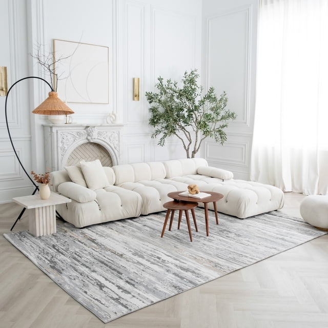 Rug Branch Atlas Camo Indoor Area Rug, Grey Beige, with a modern camo pattern in grey and beige tones, placed in a bright living room featuring a cream tufted sectional sofa, wooden nesting coffee tables, a white marble side table with dried flowers, a woven floor lamp, and a white decorative fireplace with molding and a green plant.