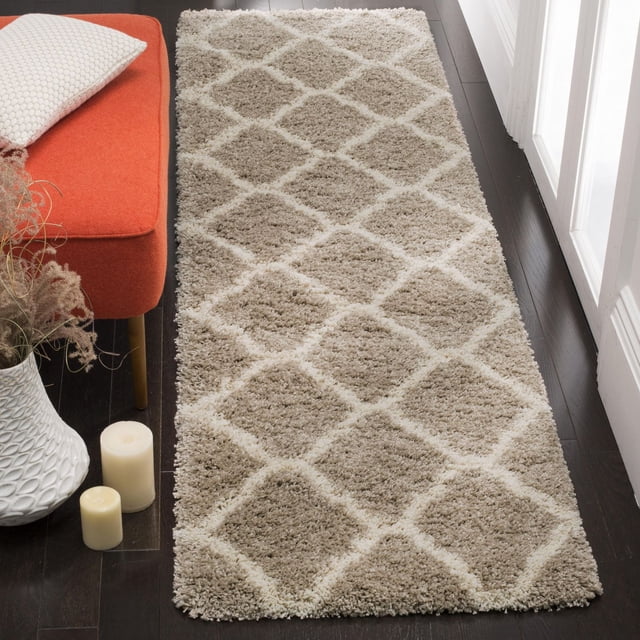 Safavieh Hudson Tapis Shag Géométrique Ligne a Mettant en Vedette un Motif de Carreaux Marocains beige et Crème en Losanges avec une pile Peluche Douce et durable Faite de Fils Synthétiques Tissés Mécaniquement, Exposé sur un Plancher de Bois Franc Foncé à Côté d'Un Banc orange avec un Oreiller Blanc, un vase Blanc avec des Plantes Séchées et Deux Bougies Crème, Idéal comme Tapis Coureur dans les Couloirs Ou les Entrées.